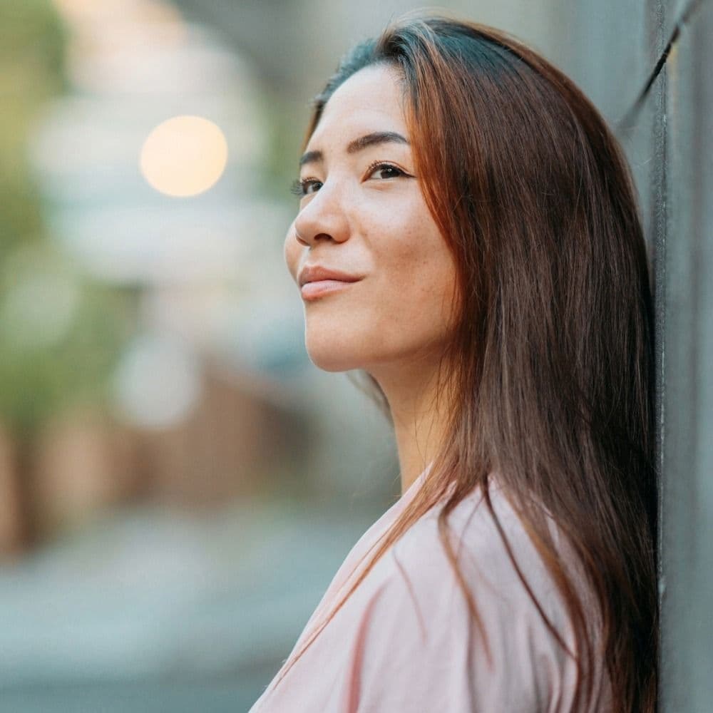 NAD+ Therapy now available at Revive Med Spa. Woman with long, straight brown hair in a pink shirt softly smiling and gazing off camera.